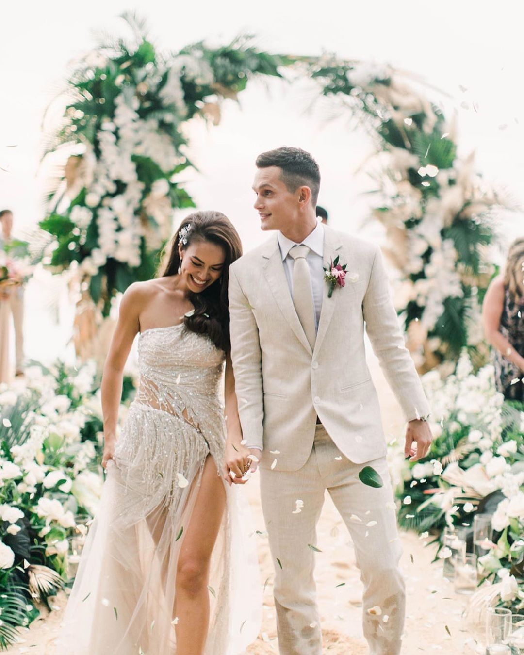 Thailand wedding with a palm frond and white flower arch, sexy glittery wedding dress and tan groom suit
